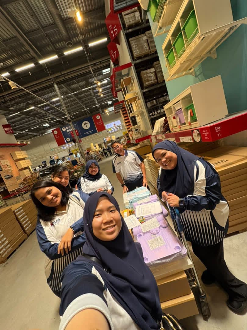 Committee members smiling beside a loaded trolley in the IKEA warehouse aisles